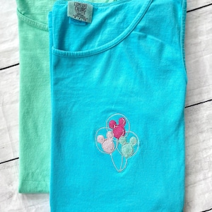 May include: Two sleeveless t-shirts, one mint green and one turquoise, are displayed. The turquoise shirt features an embroidered design of Mickey Mouse balloons. Both shirts have the "Comfort Colors" label.