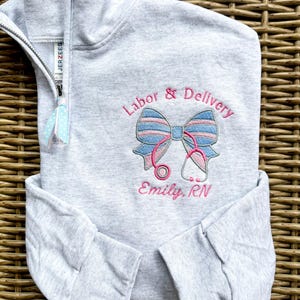 May include: A light gray quarter-zip sweatshirt with the words "Labor & Delivery" embroidered in pink above a blue and pink bow and stethoscope design. The name "Emily, RN" is embroidered below the design. The zipper has a blue and white polka dot ribbon attached.