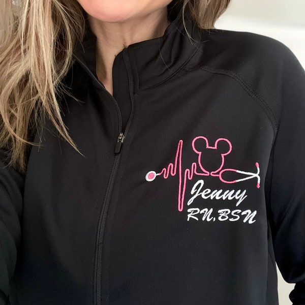 May include: A black long-sleeve zip-up jacket featuring an embroidered pink design of a heartbeat line, a mouse ear silhouette, and a stethoscope. Below the design, the text reads "Jenny" in a script font and "RN, BSN" in a block font.