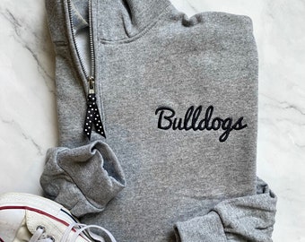 Może przedstawiać: Szara bluza z zamkiem błyskawicznym na ¼ długości i haftowanym czarnym logo "Bulldogs". Bluza leży płasko na białym marmurowym tle z parą białych tenisówek z płótna.