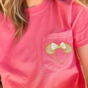 Op de afbeelding: Een roze T-shirt met een gouden geborduurde Mickey Mouse oren met een witte strik op een zak.