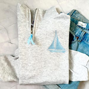 May include: A light gray sweatshirt with a quarter-zip neckline and a light blue sailboat design. The sweatshirt is paired with a pair of blue denim jeans. The jeans have a button closure and a size tag.