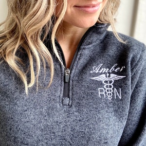 May include: A charcoal gray heathered quarter-zip pullover featuring white embroidery on the left chest. The embroidery includes the name Amber above a caduceus medical symbol and the letters RN, indicating a registered nurse professional.