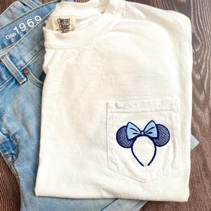 Op de afbeelding: Wit T-shirt met een blauw en wit geborduurd Minnie Mouse oren ontwerp op een zak. Het T-shirt heeft een label met de tekst "Comfort Colors".