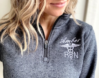 May include: A charcoal gray heathered quarter-zip pullover featuring white embroidery on the left chest. The embroidery includes the name Amber above a caduceus medical symbol and the letters RN, indicating a registered nurse professional.