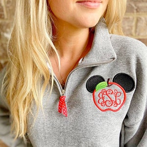Op de afbeelding: Grijs sweatshirt met een rode en witte rits en een rode appel met groene bladeren en zwarte Mickey Mouse oren geborduurd op de voorkant. De appel heeft de letters "ESL" geborduurd in de binnenkant.