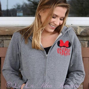 Puede incluir: Una sudadera gris con cremallera y capucha con un lazo rojo y un monograma "G" en la parte delantera. La sudadera la lleva puesta una persona con el pelo largo y rubio.