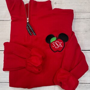 Op de afbeelding: Rood kwart-rits trui met een zwarte rits en een zwart-witte geblokte rits trekkoord. De trui heeft een geborduurde appel met een groen blad en Mickey Mouse oren, met de letters "SSK" in de appel.