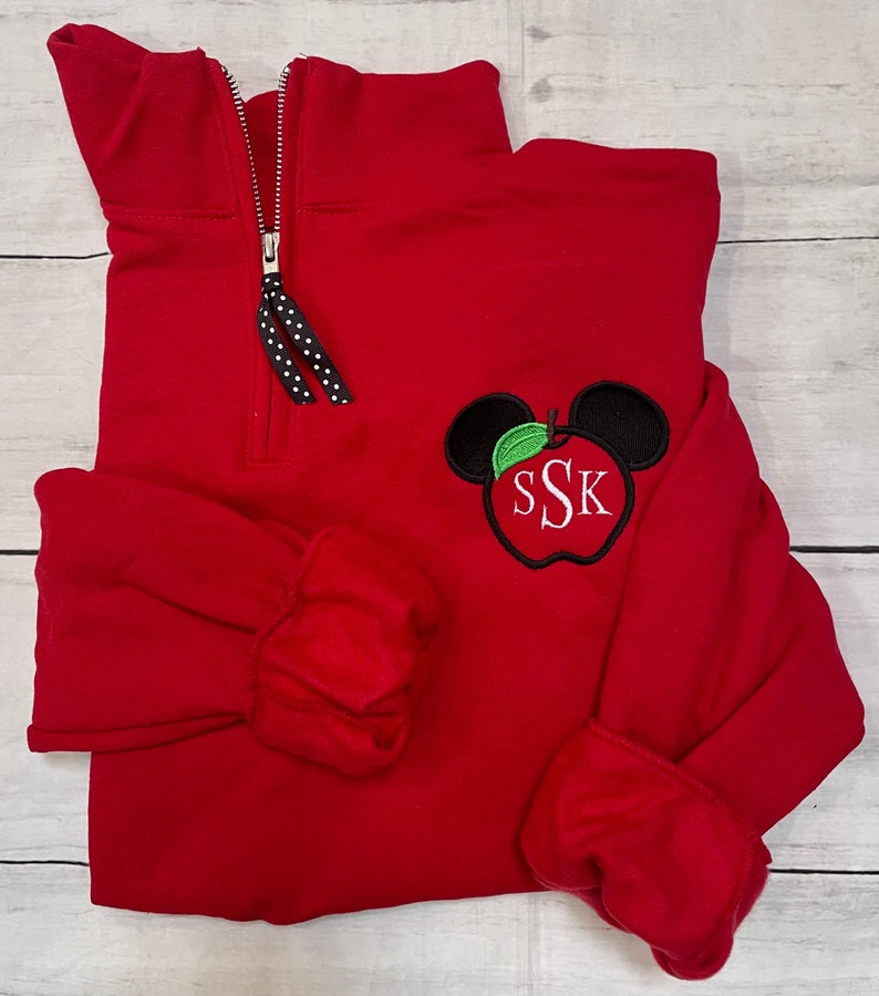 Op de afbeelding: Rood kwart-rits trui met een zwarte rits en een zwart-witte geblokte rits trekkoord. De trui heeft een geborduurde appel met een groen blad en Mickey Mouse oren, met de letters "SSK" in de appel.