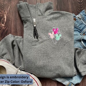 Może przedstawiać: Szara bluza z zamkiem na ćwierć z haftowanymi balonami Myszki Miki w kolorze r&oacute;żowym, zielonym i białym. Tekst "Design is embroidery Quarter Zip Color: Oxford" znajduje się na dole obrazu.