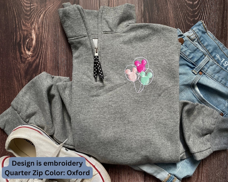 Może przedstawiać: Szara bluza z zamkiem na ćwierć z haftowanymi balonami Myszki Miki w kolorze r&oacute;żowym, zielonym i białym. Tekst "Design is embroidery Quarter Zip Color: Oxford" znajduje się na dole obrazu.