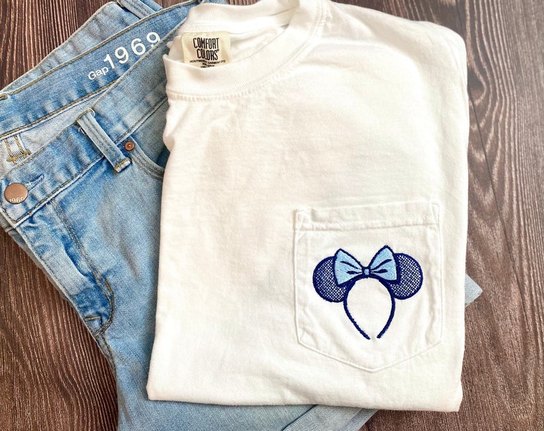 Op de afbeelding: Wit T-shirt met een blauw en wit geborduurd Minnie Mouse oren ontwerp op een zak. Het T-shirt heeft een label met de tekst "Comfort Colors".