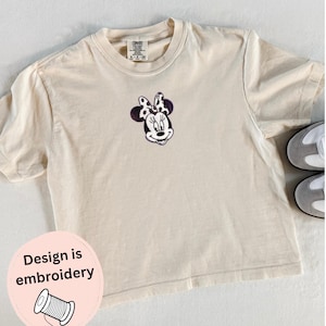 Peut inclure: T-shirt cr&egrave;me avec un motif Minnie Mouse brod&eacute;. Le t-shirt a un col rond et des manches courtes. L'&eacute;tiquette indique "Comfort Colors". Un autocollant rose avec le texte "Design is embroidery" est en bas &agrave; gauche.