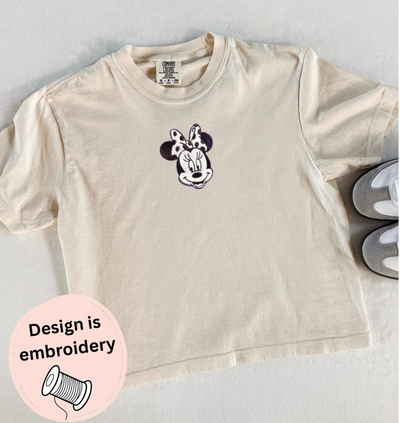 Peut inclure: T-shirt cr&egrave;me avec un motif Minnie Mouse brod&eacute;. Le t-shirt a un col rond et des manches courtes. L'&eacute;tiquette indique "Comfort Colors". Un autocollant rose avec le texte "Design is embroidery" est en bas &agrave; gauche.