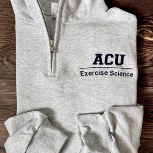 Peut inclure: Un sweat-shirt gris clair à fermeture éclair quart de zip avec le texte "ACU Exercise Science" brodé en violet foncé. Le sweat-shirt a un col et des poignets pliés, et une fermeture éclair argentée. Le vêtement est posé à plat sur une surface en bois.
