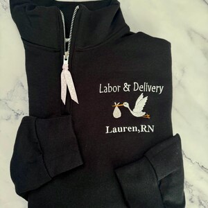 Puede incluir: Sudadera negra con cremallera de cuarto de cremallera, texto bordado en blanco y gr&aacute;fico de cig&uuml;e&ntilde;a. El texto dice "Labor & Delivery" encima del nombre "Lauren, RN". La cremallera tiene una cinta rosa y blanca con lunares.