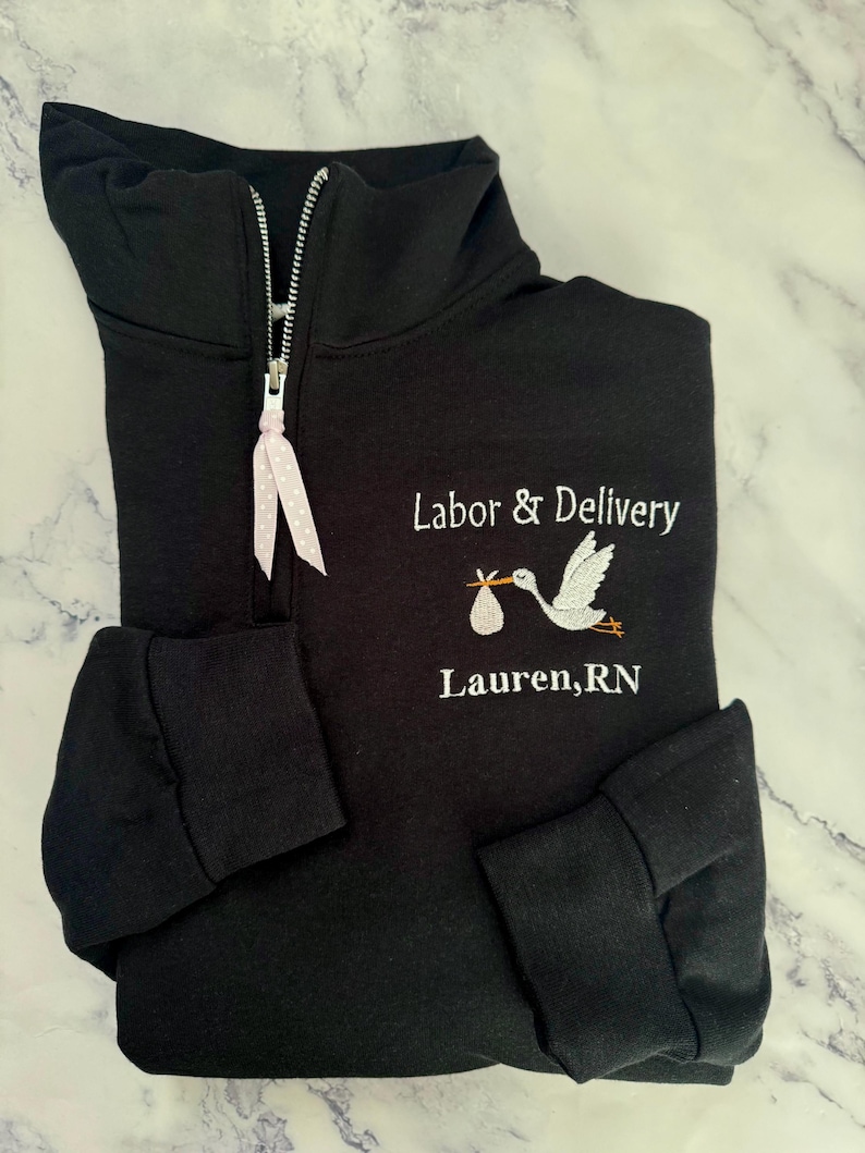 Puede incluir: Sudadera negra con cremallera de cuarto de cremallera, texto bordado en blanco y gr&aacute;fico de cig&uuml;e&ntilde;a. El texto dice "Labor & Delivery" encima del nombre "Lauren, RN". La cremallera tiene una cinta rosa y blanca con lunares.