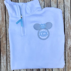Pu&ograve; includere: Felpa bianca con zip a un quarto con un designo ricamato grigio a forma di orecchio di Topolino con un anello di diamanti blu. La felpa ha anche un monogramma ricamato grigio con le lettere "JBM".