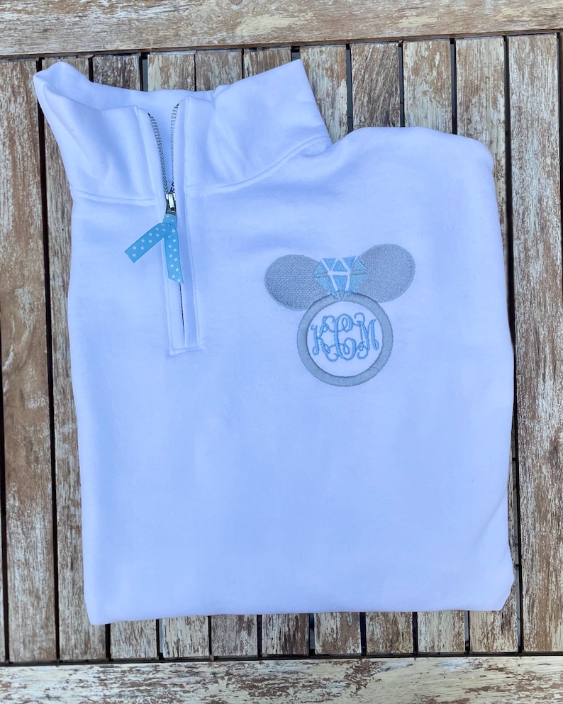 Pu&ograve; includere: Felpa bianca con zip a un quarto con un designo ricamato grigio a forma di orecchio di Topolino con un anello di diamanti blu. La felpa ha anche un monogramma ricamato grigio con le lettere "JBM".