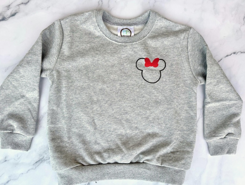 Op de afbeelding: Een lichtgrijs sweatshirt met een zwarte omtrek van een muizenkop en een rode strik. De top met lange mouwen en ronde hals heeft geribde manchetten en zoom. Het sweatshirt is gemaakt van een zacht materiaal.
