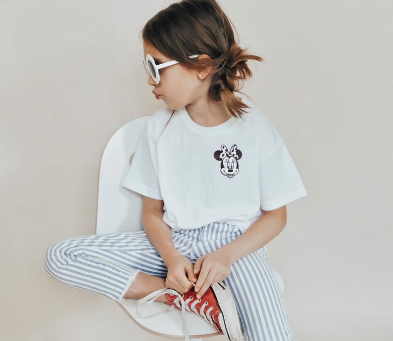 May include: A white t-shirt with a Minnie Mouse graphic, paired with blue and white striped pants and red sneakers. The child is wearing white sunglasses and sitting on a white chair.