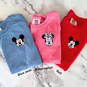 May include: Three Comfort Colors t-shirts in blue jean, watermelon, and red. Each shirt features an embroidered cartoon character face. The blue jean shirt has Mickey Mouse, the watermelon shirt has Minnie Mouse, and the red shirt has Mickey Mouse.