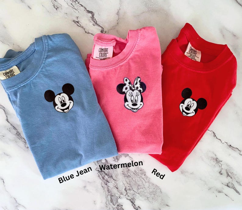 May include: Three Comfort Colors t-shirts in blue jean, watermelon, and red. Each shirt features an embroidered cartoon character face. The blue jean shirt has Mickey Mouse, the watermelon shirt has Minnie Mouse, and the red shirt has Mickey Mouse.