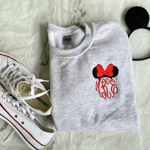 Puede incluir: Una sudadera gris con un lazo rojo y orejas negras bordadas en la parte delantera. La sudadera tiene las letras "MKE" bordadas en rojo debajo del lazo. Tambi&eacute;n se ve un par de zapatillas Converse blancas en la imagen.