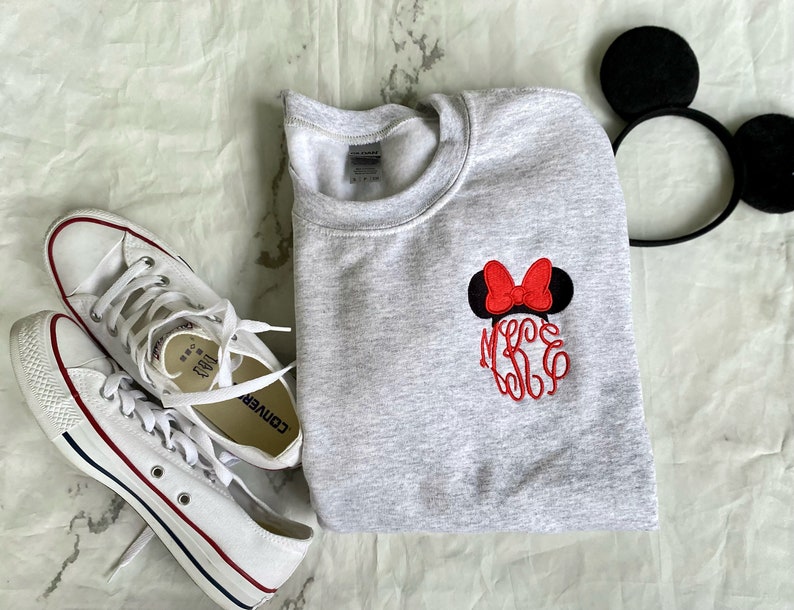 Puede incluir: Una sudadera gris con un lazo rojo y orejas negras bordadas en la parte delantera. La sudadera tiene las letras "MKE" bordadas en rojo debajo del lazo. Tambi&eacute;n se ve un par de zapatillas Converse blancas en la imagen.