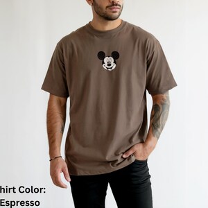 May include: An espresso brown short-sleeved t-shirt featuring a centered graphic of the Mickey Mouse character head. The design shows the iconic black ears and facial features of the character printed on the chest of the solid brown garment.