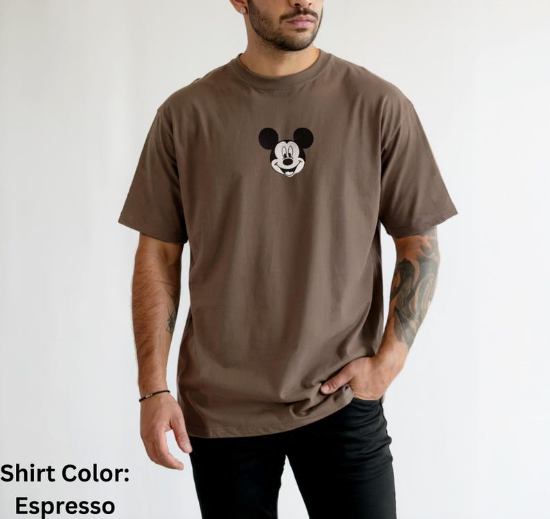 May include: An espresso brown short-sleeved t-shirt featuring a centered graphic of the Mickey Mouse character head. The design shows the iconic black ears and facial features of the character printed on the chest of the solid brown garment.
