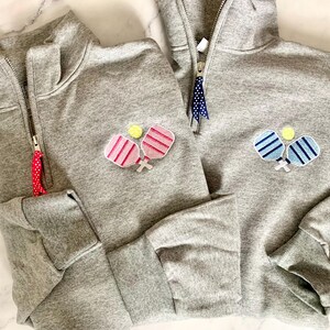 May include: Two grey sweatshirts with a half-zip fastening. One sweatshirt features a pink and white paddleball design, and the other has a blue and white paddleball design.