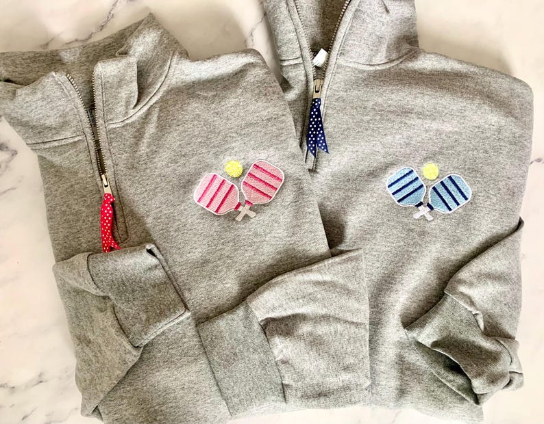 May include: Two grey sweatshirts with a half-zip fastening. One sweatshirt features a pink and white paddleball design, and the other has a blue and white paddleball design.