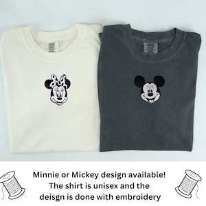 May include: Two crew neck t-shirts, one off-white and one charcoal gray, each featuring a small embroidered character face on the chest. The off-white shirt displays a Minnie Mouse design, while the charcoal gray shirt features a Mickey Mouse design.