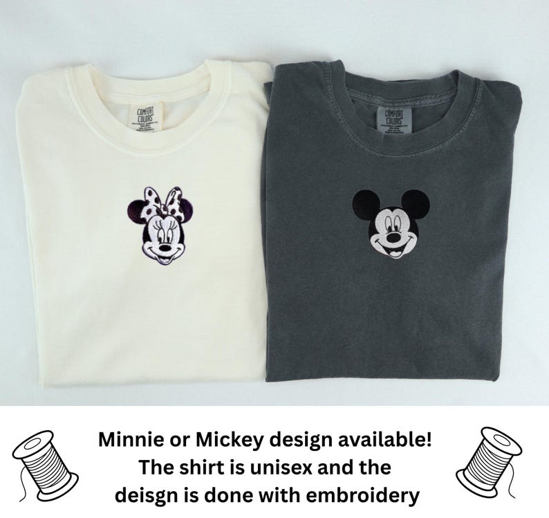 May include: Two crew neck t-shirts, one off-white and one charcoal gray, each featuring a small embroidered character face on the chest. The off-white shirt displays a Minnie Mouse design, while the charcoal gray shirt features a Mickey Mouse design.