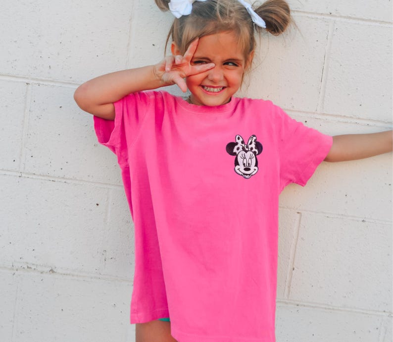 May include: A bright pink t-shirt featuring a Minnie Mouse graphic on the front. The graphic is a black and white image of Minnie Mouse's face with a polka dot bow. The shirt is worn by a young person.