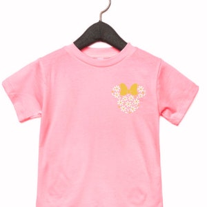 May include: Pink short-sleeved t-shirt with a floral Minnie Mouse design on the front. The design features white flowers and a yellow bow.