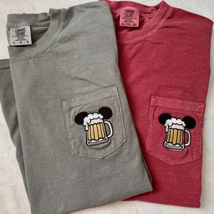 May include: A gray and a red short-sleeved t-shirt, each featuring a chest pocket with an embroidered graphic of a beer mug with foam and black mouse ears. The gray shirt has a size L tag, and the red shirt has a size XL tag, both labeled Comfort Colors.