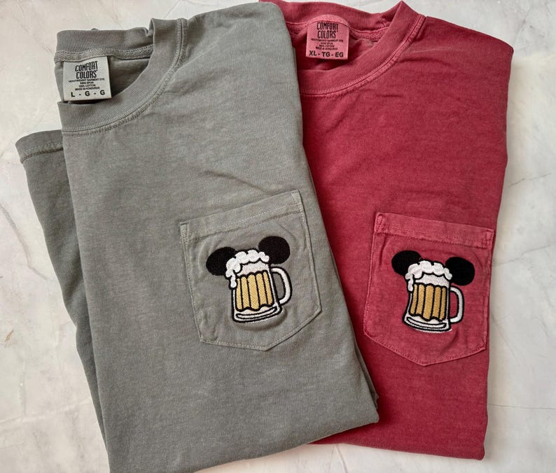 May include: A gray and a red short-sleeved t-shirt, each featuring a chest pocket with an embroidered graphic of a beer mug with foam and black mouse ears. The gray shirt has a size L tag, and the red shirt has a size XL tag, both labeled Comfort Colors.