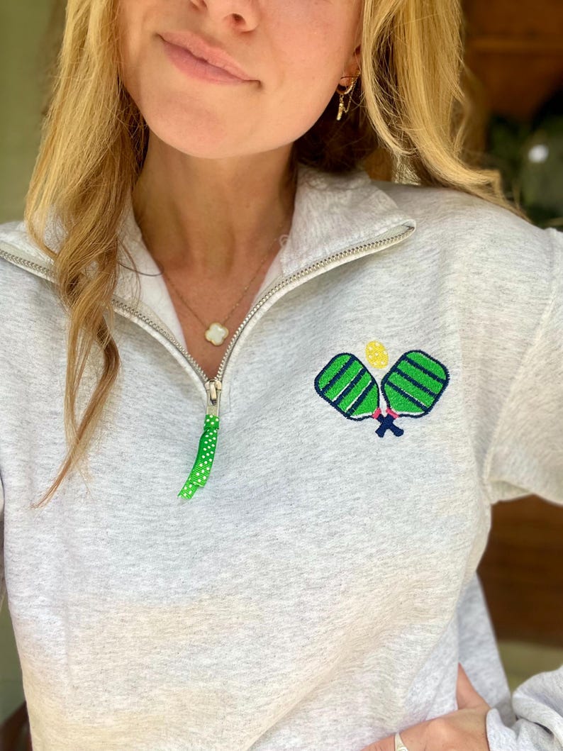 May include: A grey sweatshirt with an embroidered design of two paddleball rackets in green and yellow. The sweatshirt has a zip and a green polka dot ribbon.