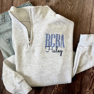May include: A light gray quarter-zip sweatshirt with blue embroidered text that reads "BCBA Haley." The sweatshirt is folded next to a pair of light blue denim jeans. The zipper is silver.