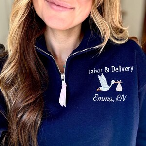 Puede incluir: Sudadera azul marino con cremallera de cuarto de cremallera y cig&uuml;e&ntilde;a bordada que lleva un beb&eacute;. El texto dice "Labor & Delivery" encima de la cig&uuml;e&ntilde;a y "Emma, RN" debajo. El tirador de la cremallera tiene una cinta rosa.