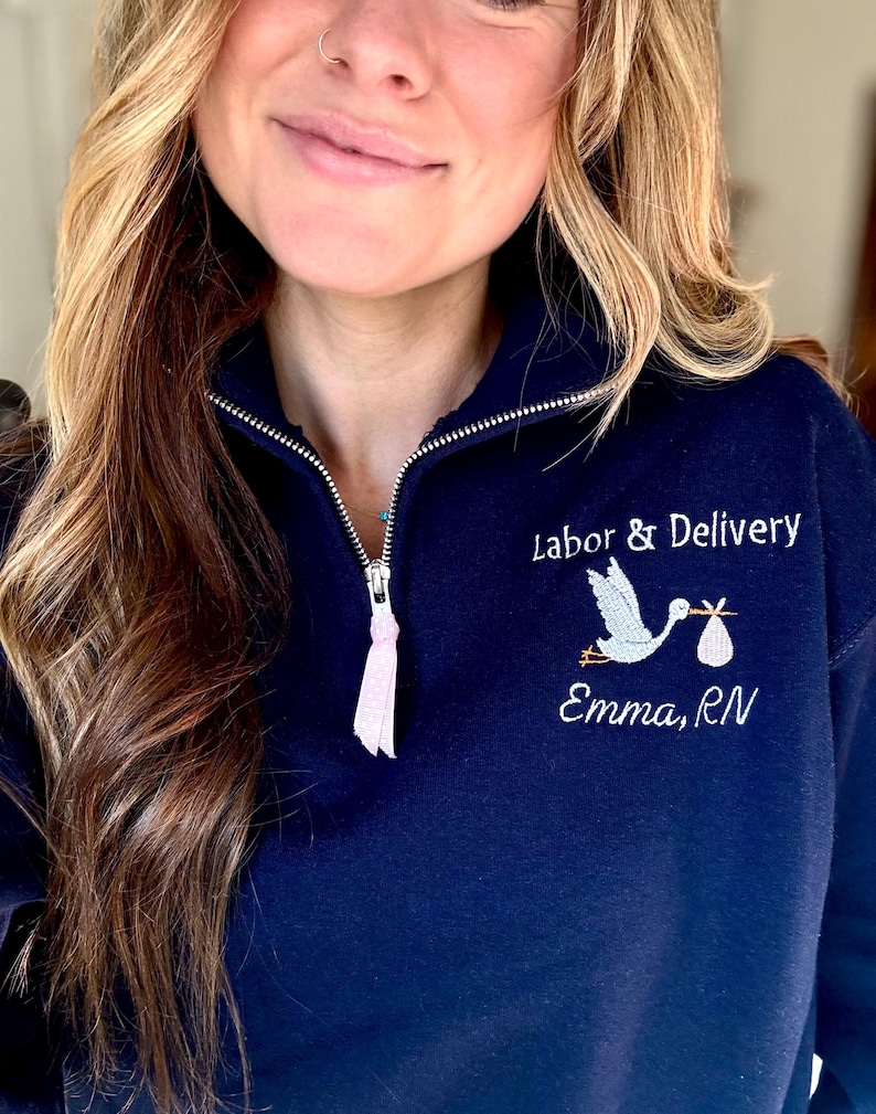 Puede incluir: Sudadera azul marino con cremallera de cuarto de cremallera y cig&uuml;e&ntilde;a bordada que lleva un beb&eacute;. El texto dice "Labor & Delivery" encima de la cig&uuml;e&ntilde;a y "Emma, RN" debajo. El tirador de la cremallera tiene una cinta rosa.