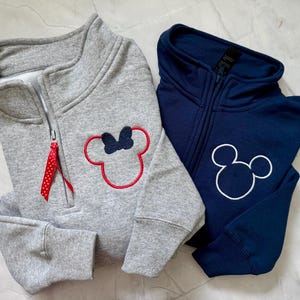 May include: Two zip-up sweatshirts. One is gray with a red and white polka dot ribbon on the zipper and a Minnie Mouse design. The other is navy blue with a Mickey Mouse design. Both have a high collar.