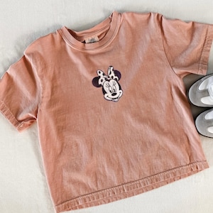 Peut inclure: T-shirt p&ecirc;che &agrave; manches courtes avec un motif brod&eacute; de Minnie Mouse sur le devant. Le motif repr&eacute;sente le visage de Minnie avec un n&oelig;ud &agrave; pois noirs et blancs. Une paire de baskets grises et blanches est visible dans le coin.