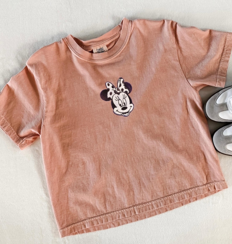 Peut inclure: T-shirt p&ecirc;che &agrave; manches courtes avec un motif brod&eacute; de Minnie Mouse sur le devant. Le motif repr&eacute;sente le visage de Minnie avec un n&oelig;ud &agrave; pois noirs et blancs. Une paire de baskets grises et blanches est visible dans le coin.
