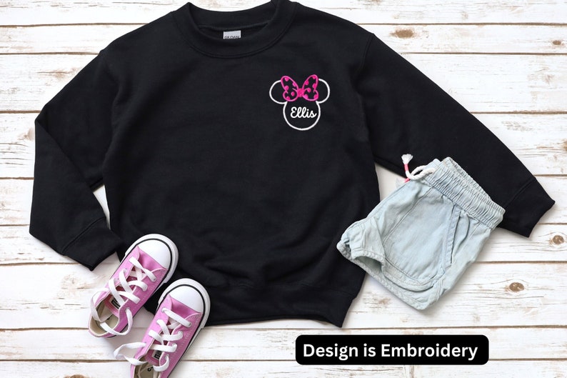 May include: Black sweatshirt with a white embroidered Minnie Mouse design with a pink bow and the name "Ellis" on a white background. The design is on the left chest of the sweatshirt.