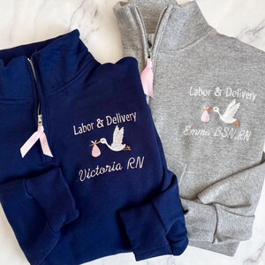 Puede incluir: Dos sudaderas con cremallera de cuarto de c&iacute;rculo, una azul marino y otra gris, bordadas con "Labor & Delivery" y una cig&uuml;e&ntilde;a que lleva un beb&eacute;. La sudadera azul marino dice "Victoria RN" y la gris dice "Emma BSN, RN". Cada una tiene una cinta rosa.