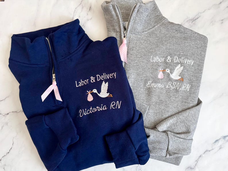 Puede incluir: Dos sudaderas con cremallera de cuarto de c&iacute;rculo, una azul marino y otra gris, bordadas con "Labor & Delivery" y una cig&uuml;e&ntilde;a que lleva un beb&eacute;. La sudadera azul marino dice "Victoria RN" y la gris dice "Emma BSN, RN". Cada una tiene una cinta rosa.