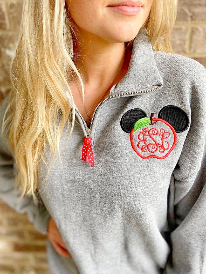 Op de afbeelding: Grijs sweatshirt met een rode en witte rits en een rode appel met groene bladeren en zwarte Mickey Mouse oren geborduurd op de voorkant. De appel heeft de letters "ESL" geborduurd in de binnenkant.